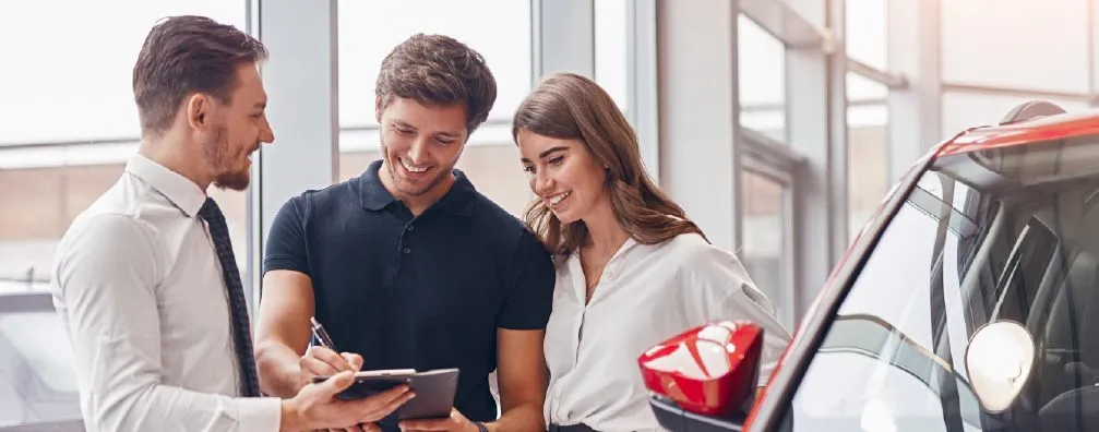 Dealership manager reviewing a tablet with a young couple beside a car