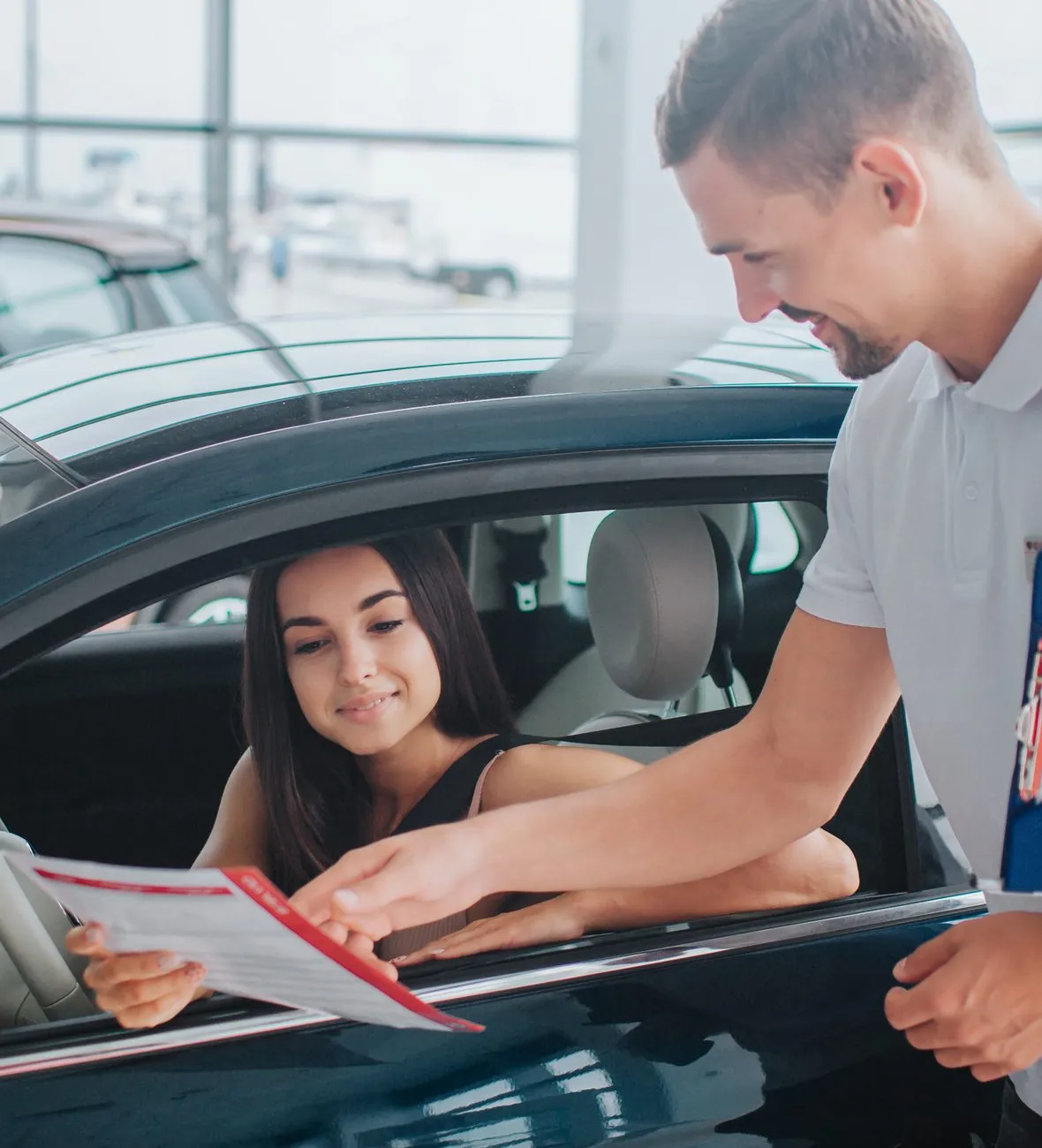 Salesperson showing a brochure to a smiling woman seated in a car at a dealership