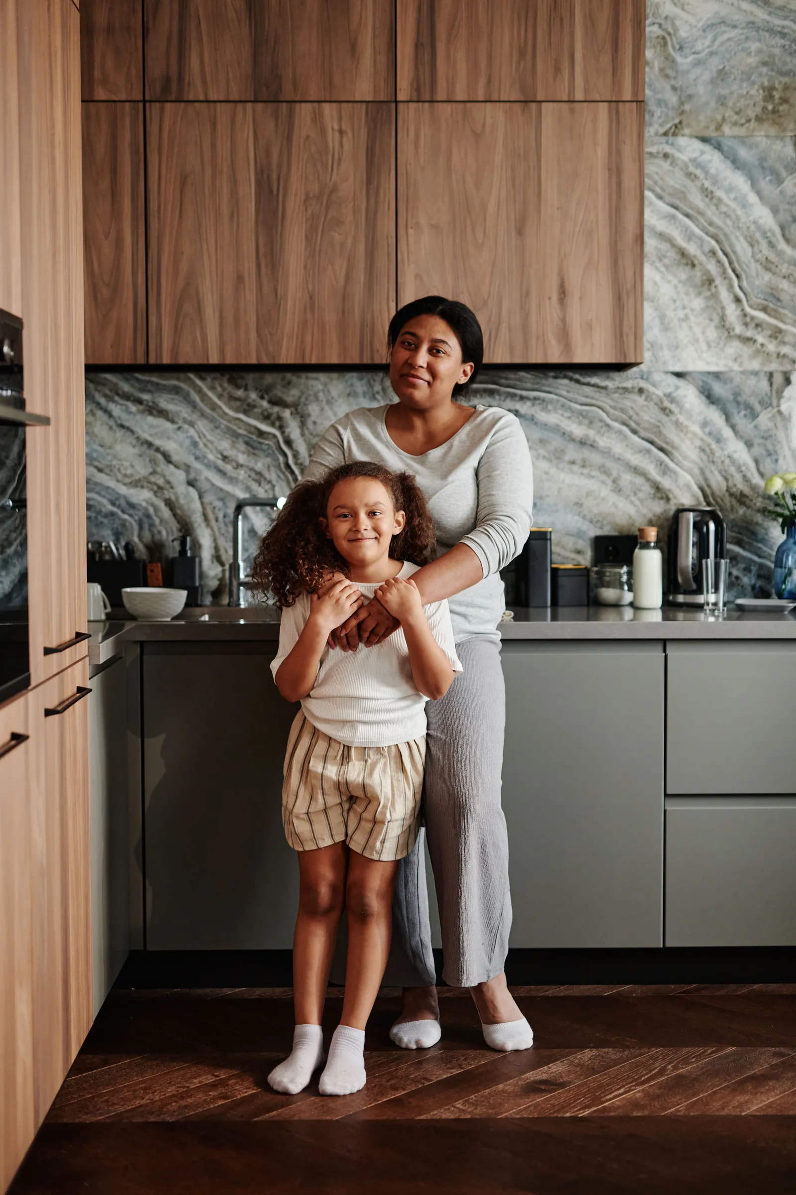 A mother and her young daughter smile together in a kitchen.