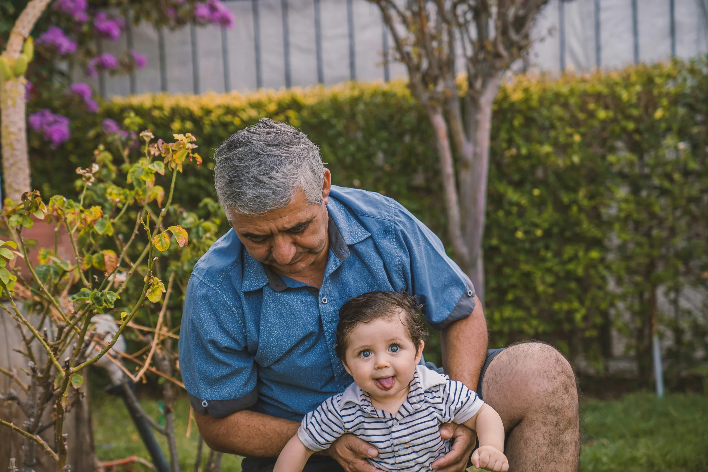 A grandfather kneels in a garden, holding a smiling baby.