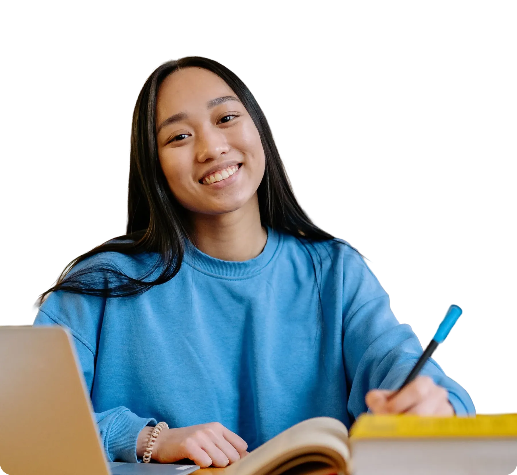 Smiling student with long dark hair and a blue sweatshirt, writing in a book at a table beside a laptop.