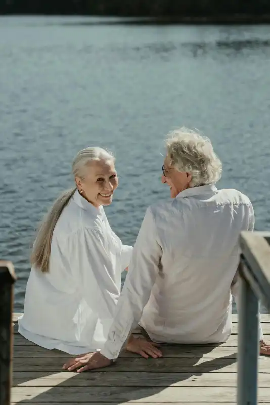 Älteres Paar sitzt lächelnd auf einem Steg am Wasser