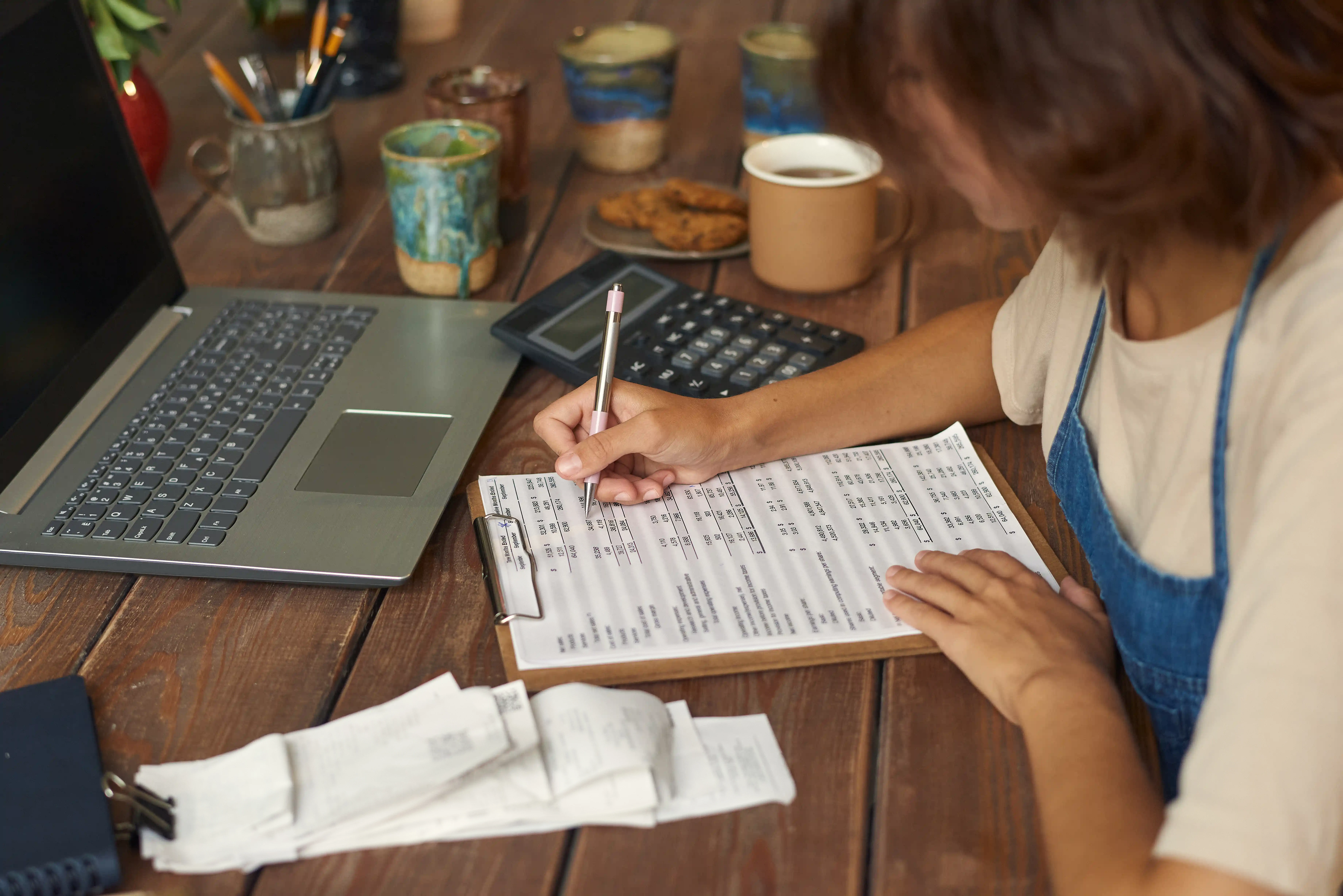Person writing on a financial worksheet on a clipboard beside a laptop, calculator, receipts, coffee cup, and cookies on a wooden table.