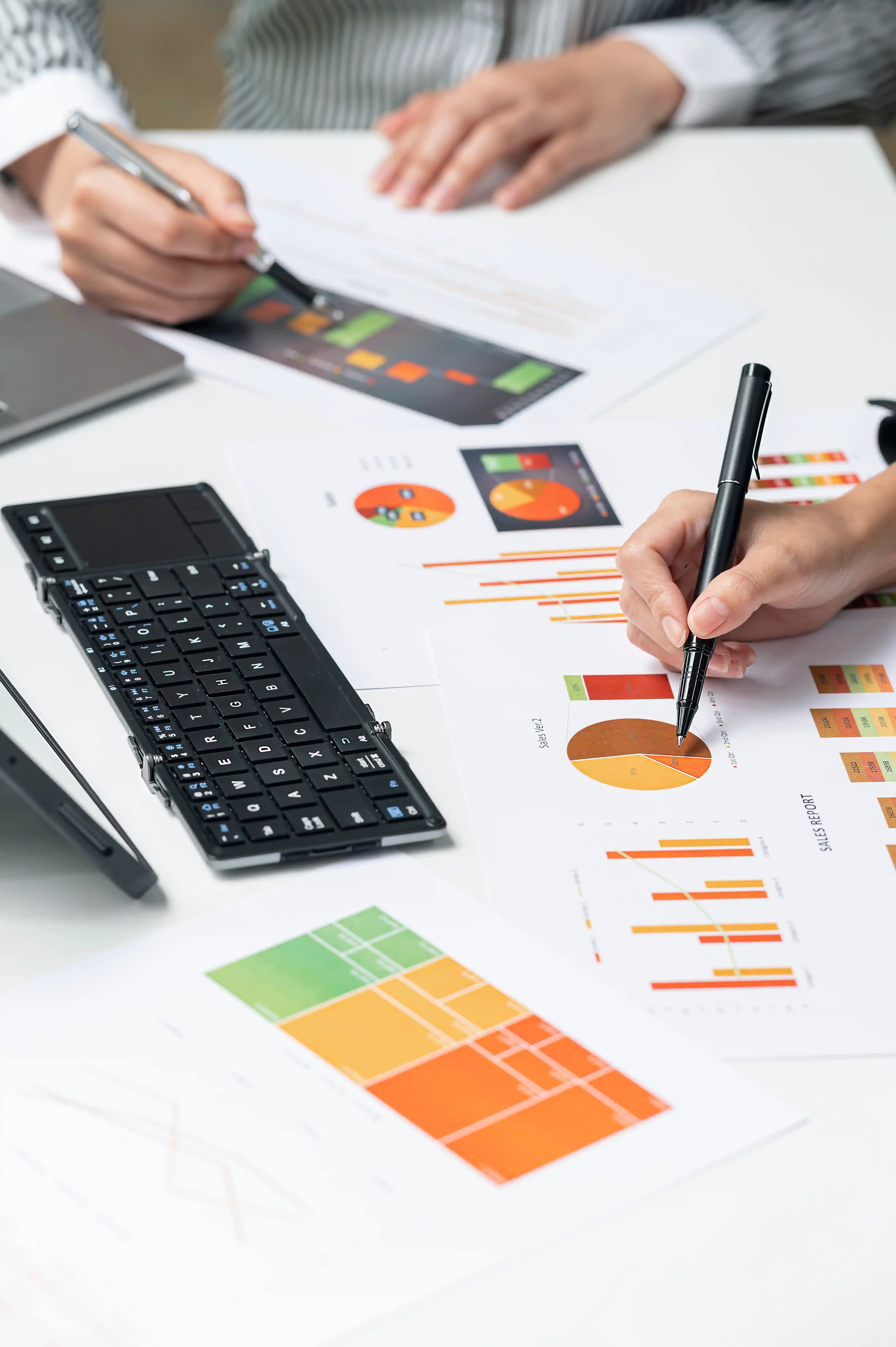 Two people reviewing and marking colorful business charts and graphs on a white desk with a foldable black keyboard nearby.