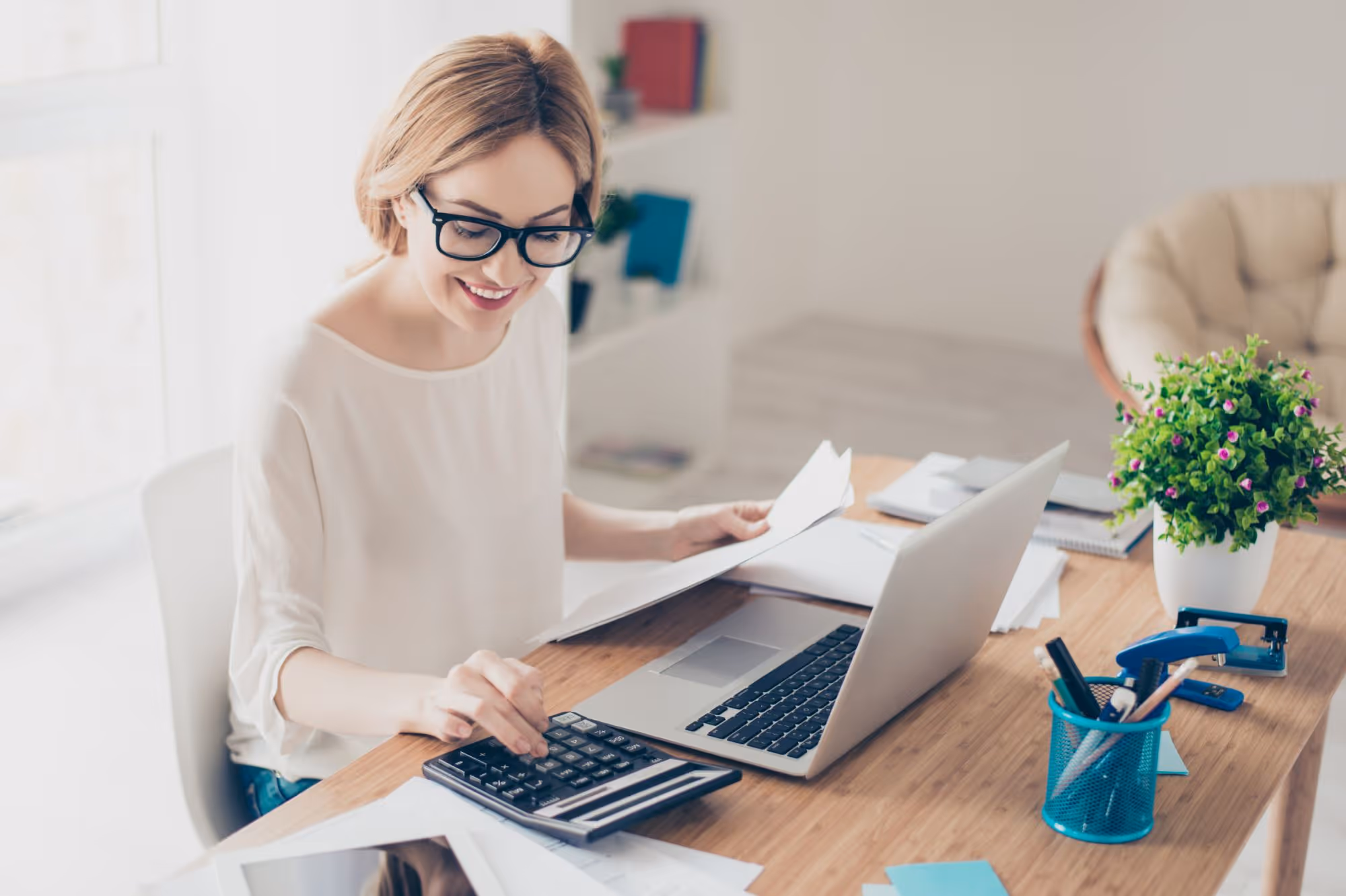 Smiling woman wearing glasses working at a desk with a calculator, laptop, and documents.