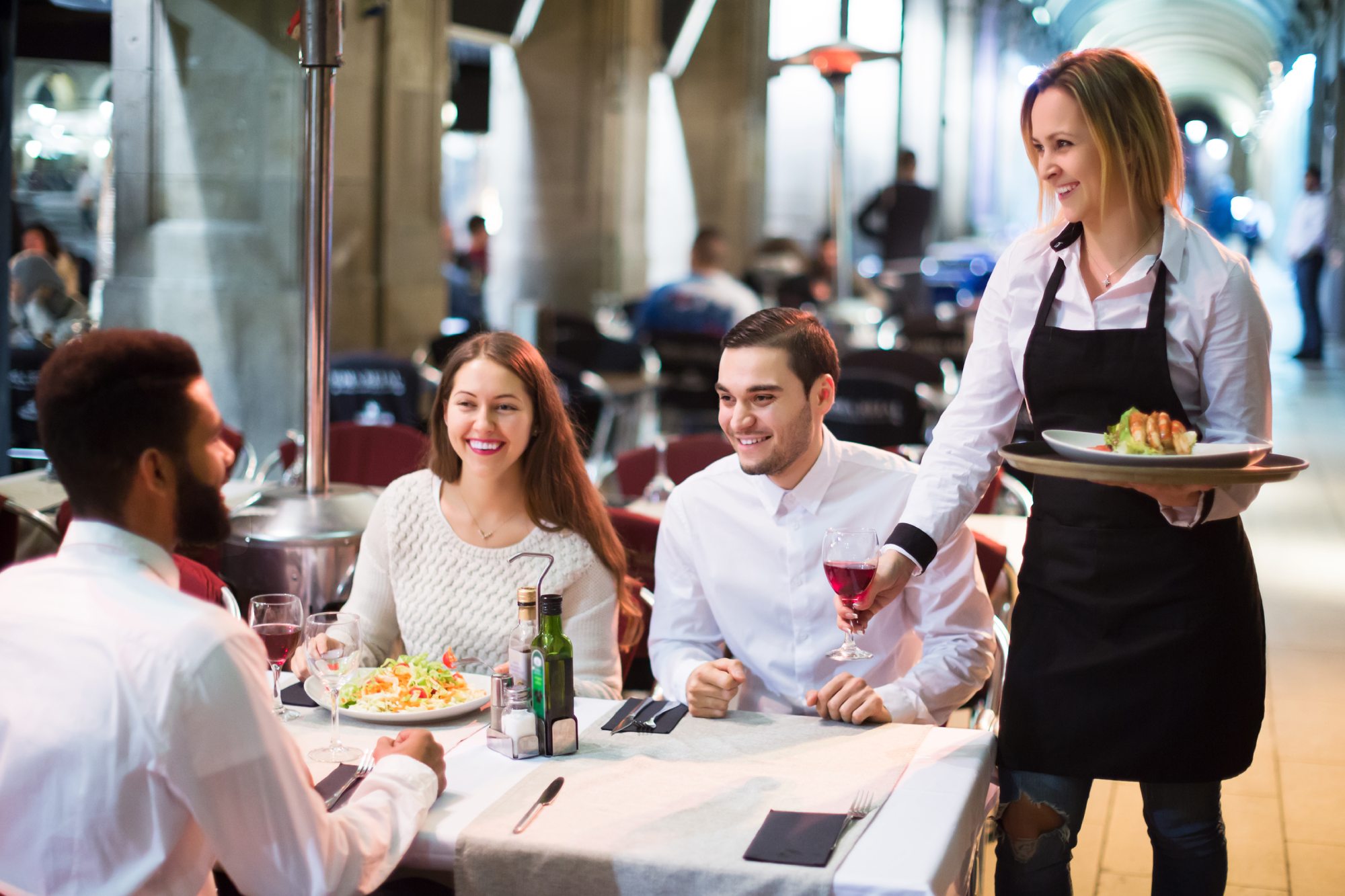 Waitress serving wine and a plate of food to three happy diners at a restaurant table.