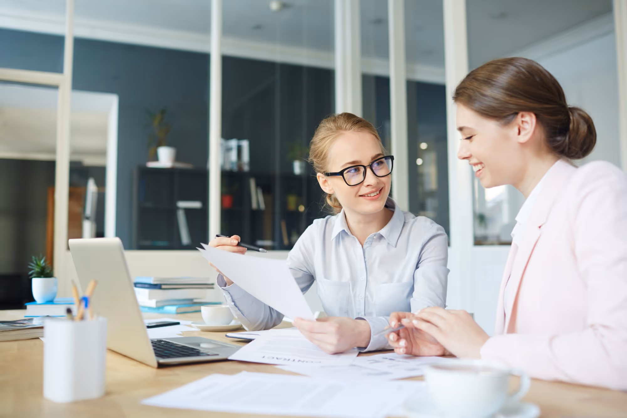 Two professional women sitting at a desk reviewing documents with a laptop and cups of coffee in an office.