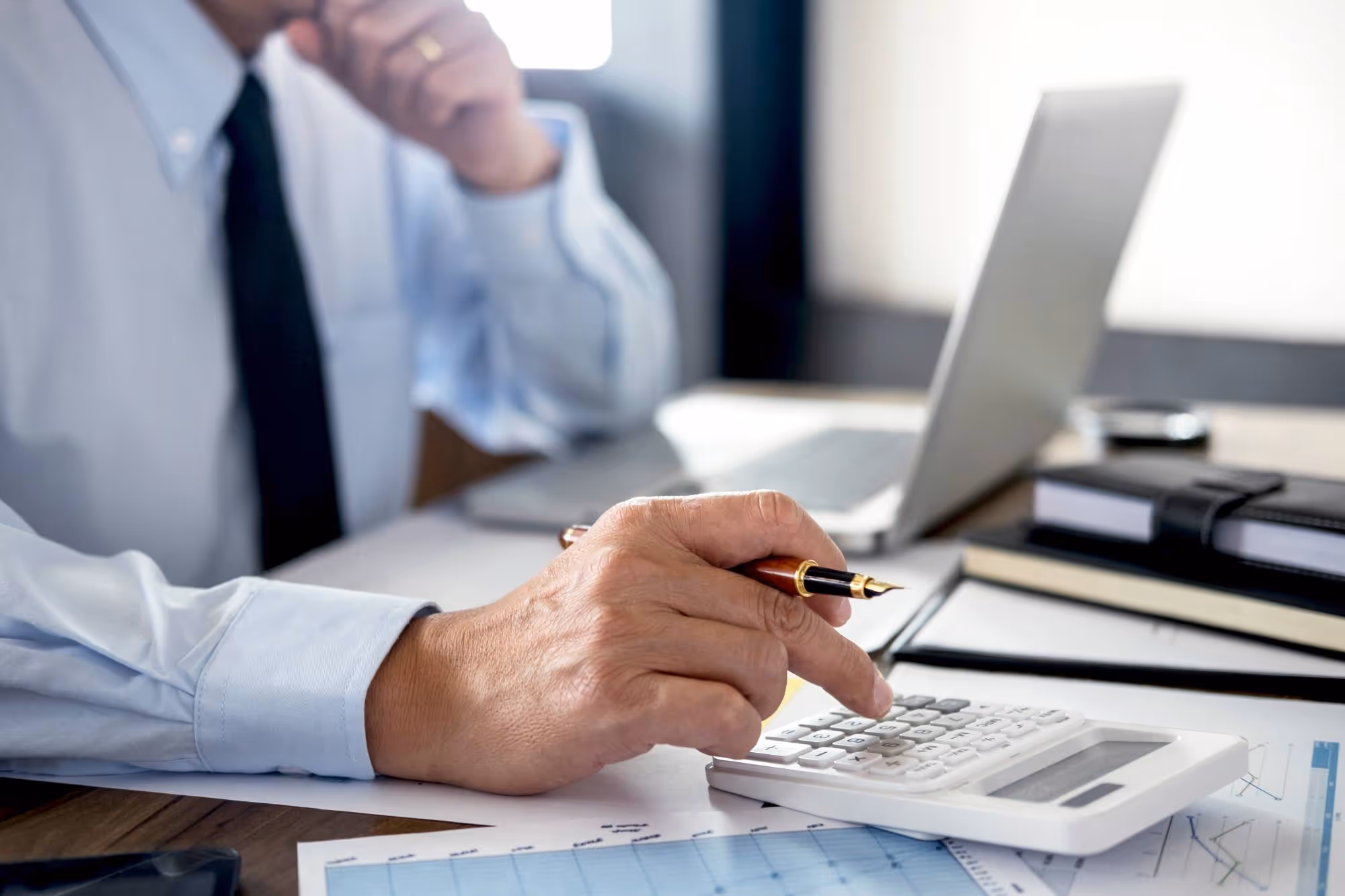 Person in business attire using a white calculator on a desk with financial documents and a laptop.