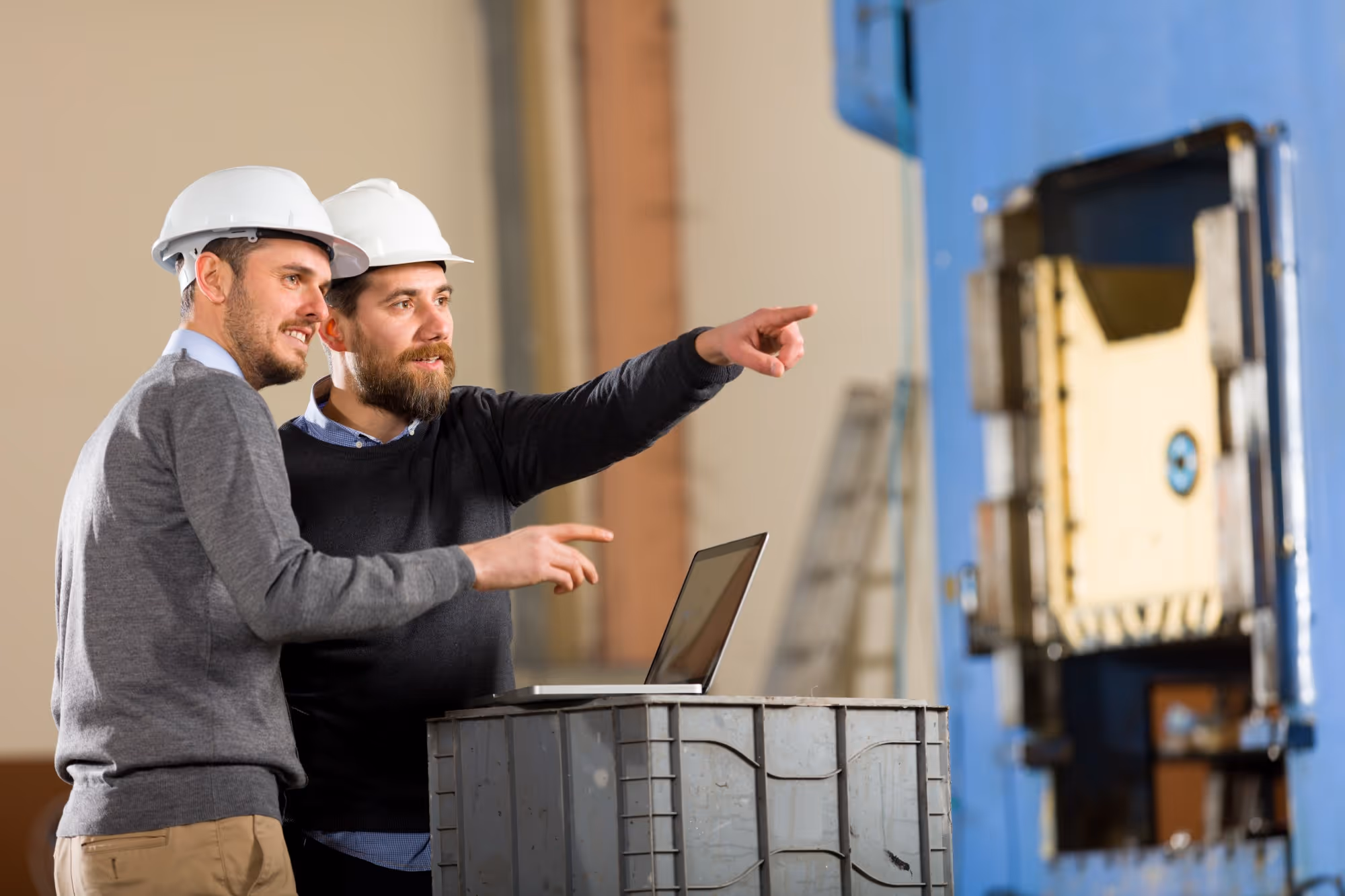 Two male engineers wearing white hard hats discussing and pointing near industrial machinery with a laptop on a metal container.