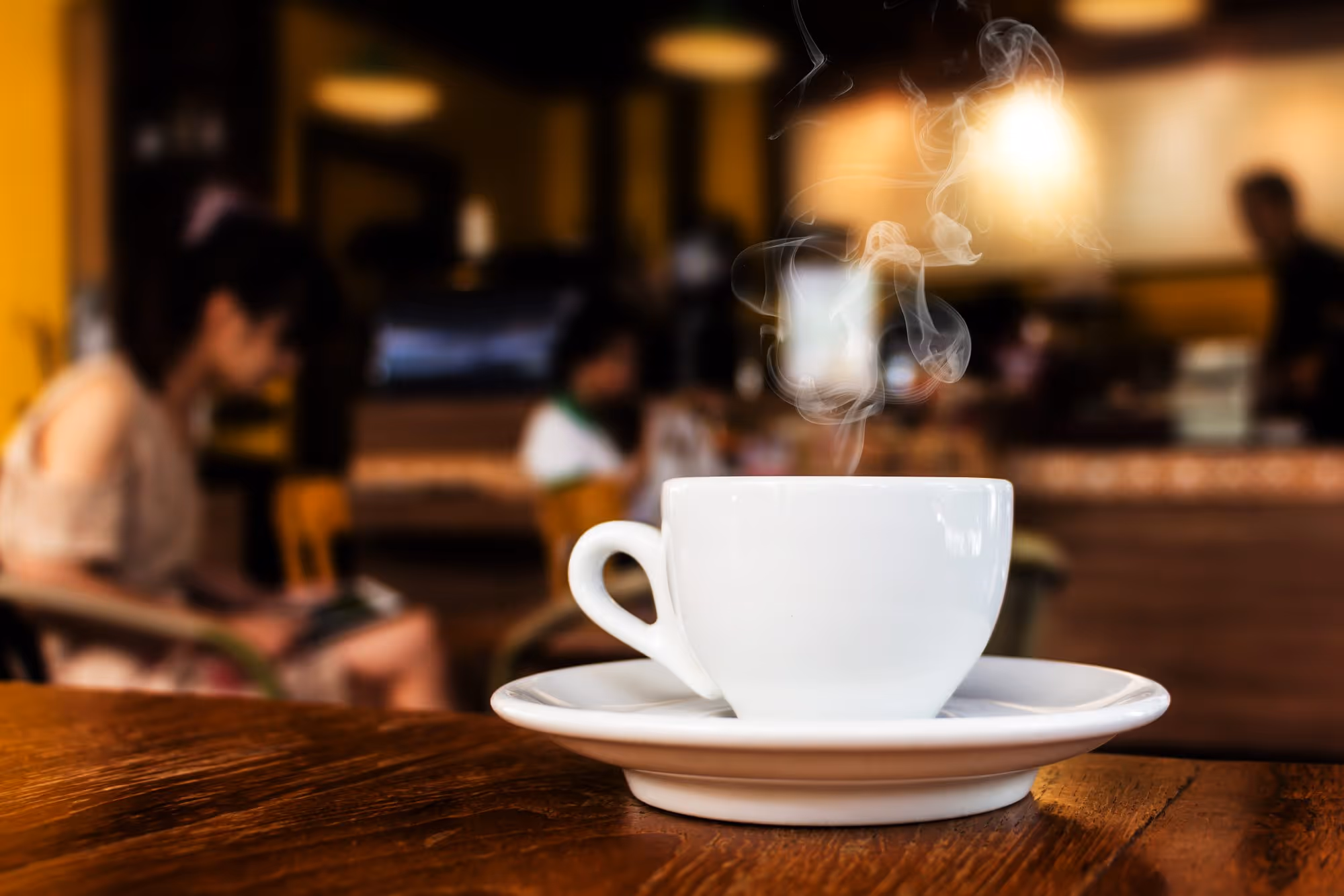White coffee cup with steam rising on a wooden table inside a cozy café with blurred people in the background.