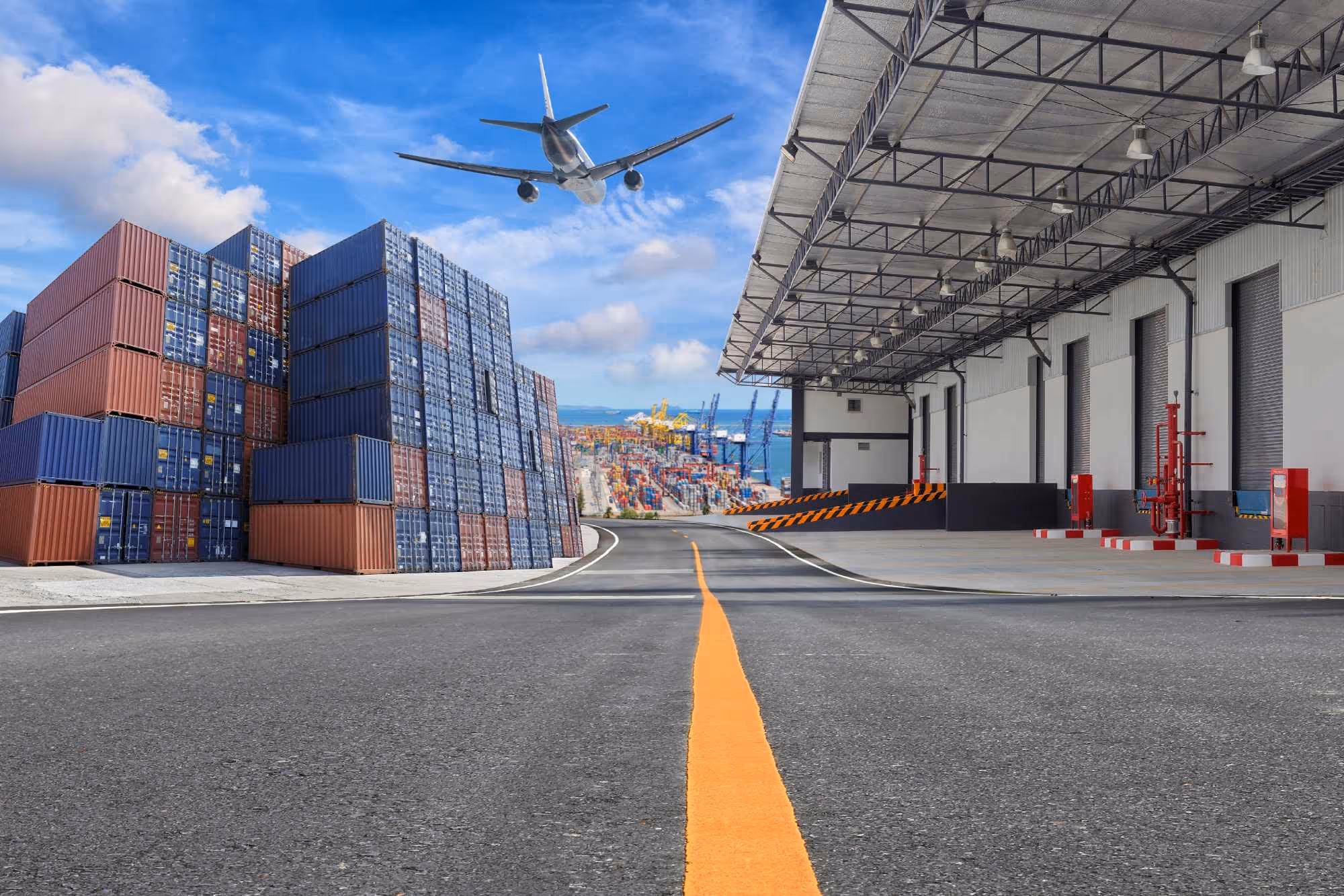 Airplane flying over a shipping port with stacked cargo containers and an industrial warehouse under a blue sky.