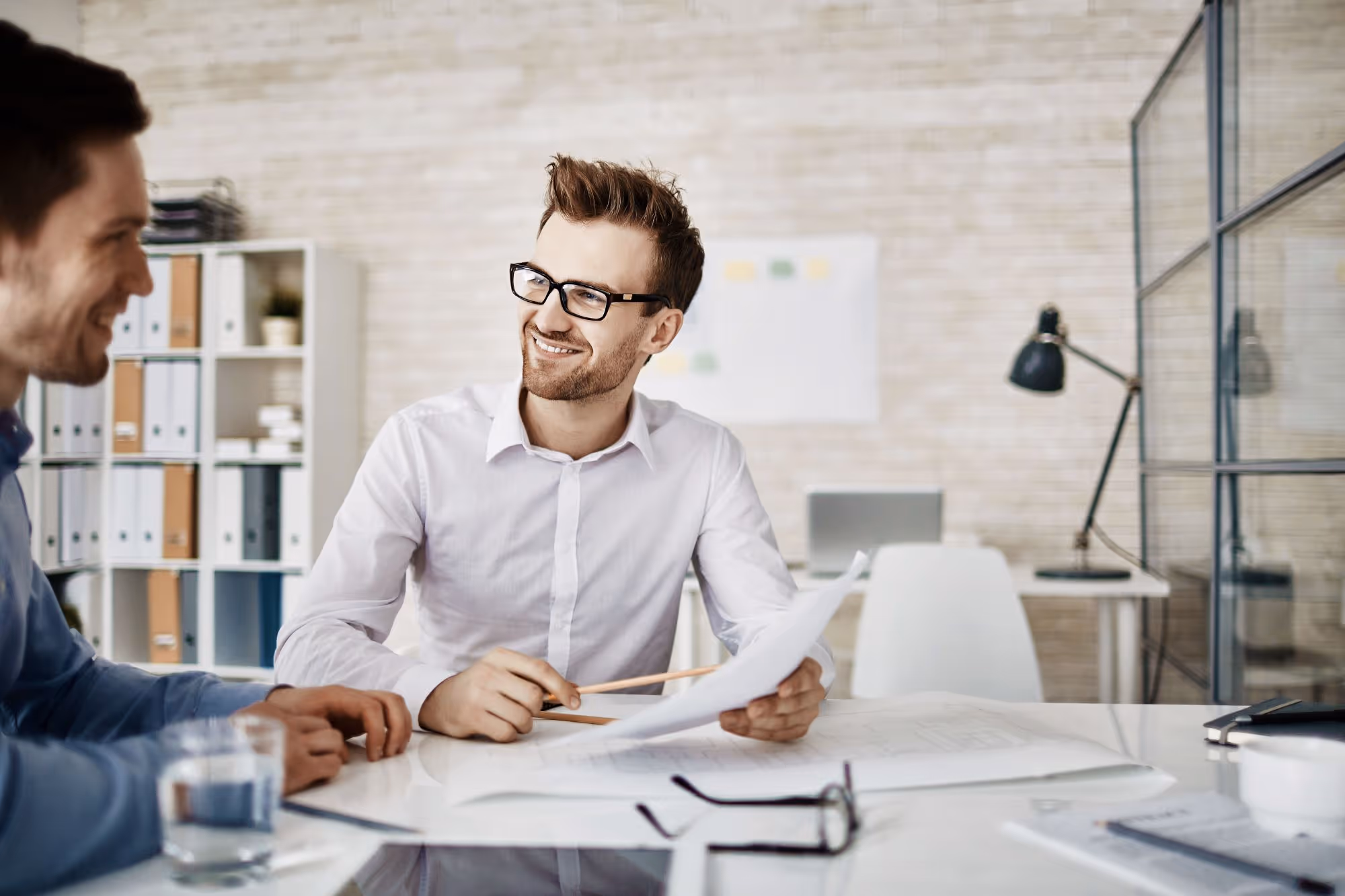 Two young men in an office having a discussion with one holding papers and a pencil.