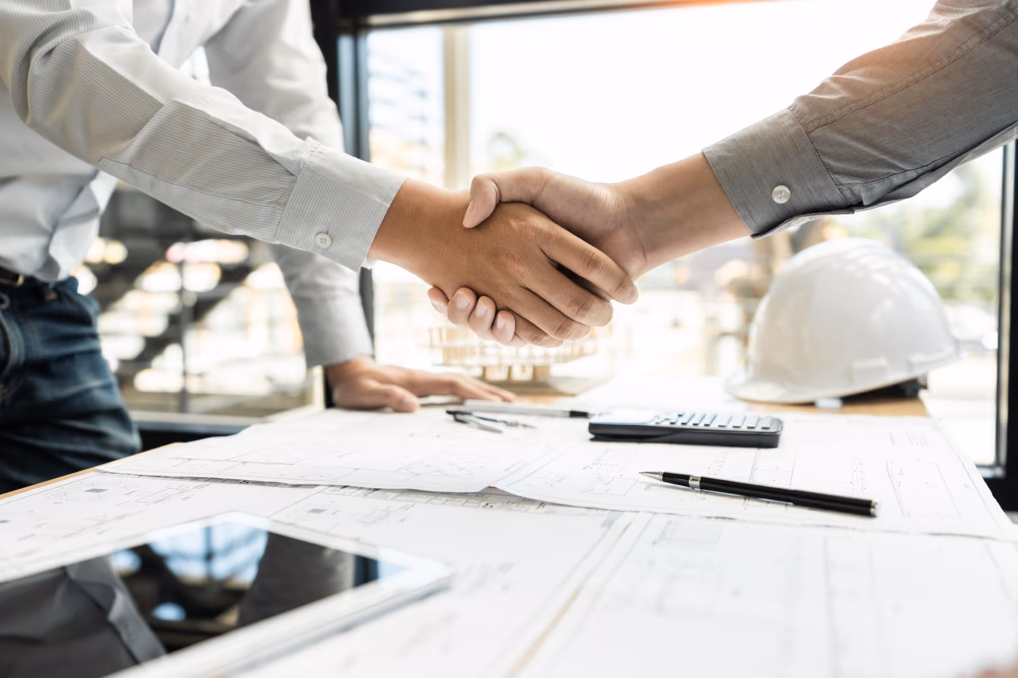 Two people in business shirts shaking hands over architectural blueprints on a table with a calculator, pen, tablet, and a white construction helmet.