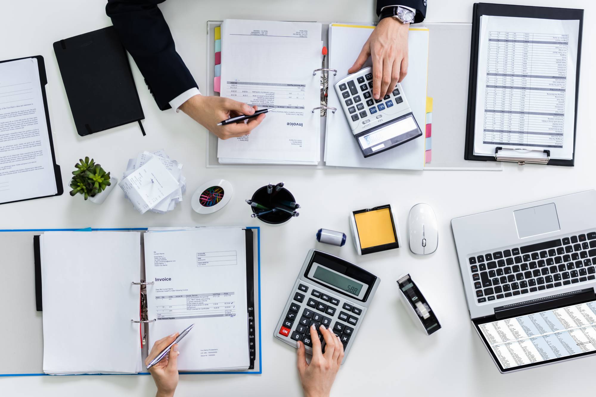 Overhead view of three people working on business invoices and calculations with calculators, documents, and a laptop on a white desk.