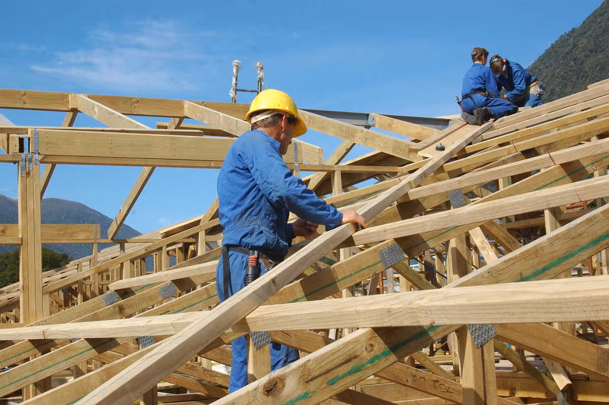 Construction workers in blue coveralls assembling wooden roof trusses under a clear blue sky.
