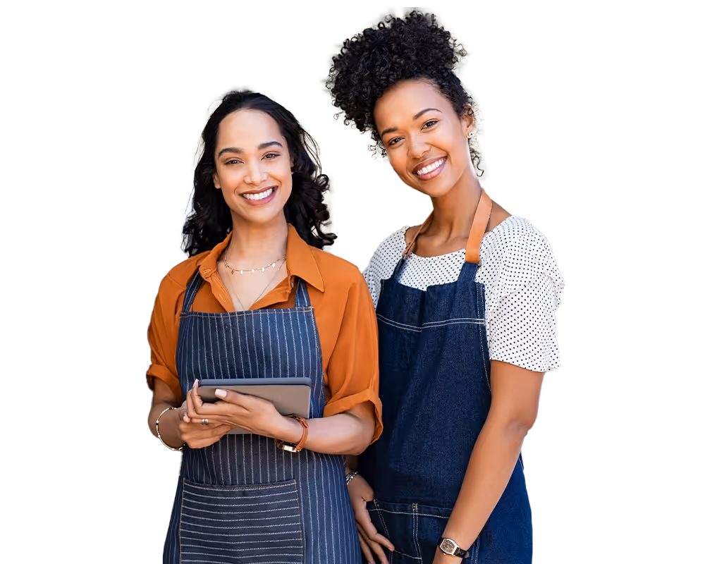 Two smiling women wearing aprons, one holding a tablet, posing together.