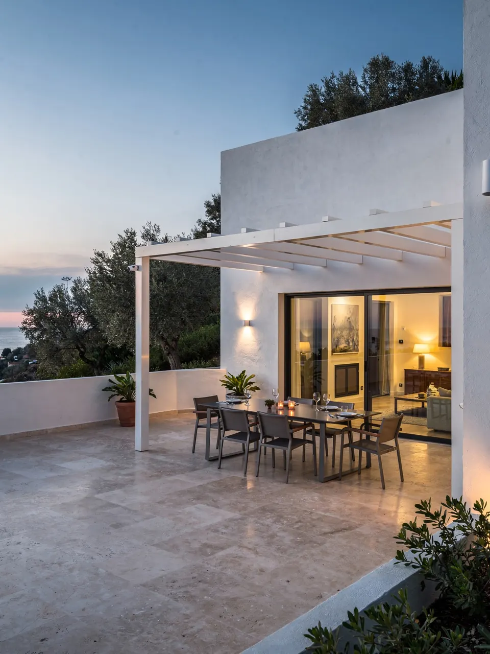 The terrace of a white villa at dusk, with a dining table under a pergola