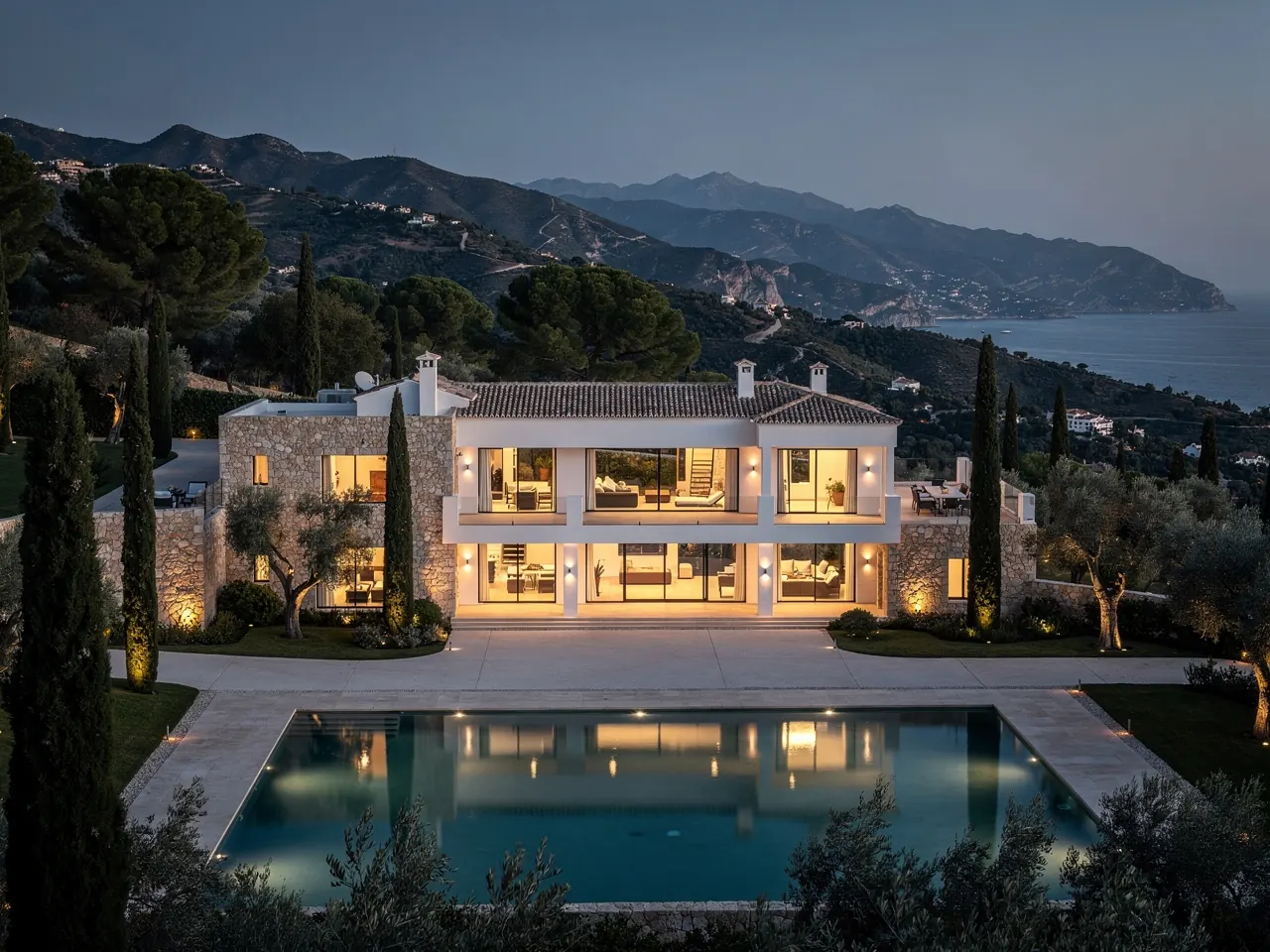 A large stone and white estate home with a long pool in the hills at dusk