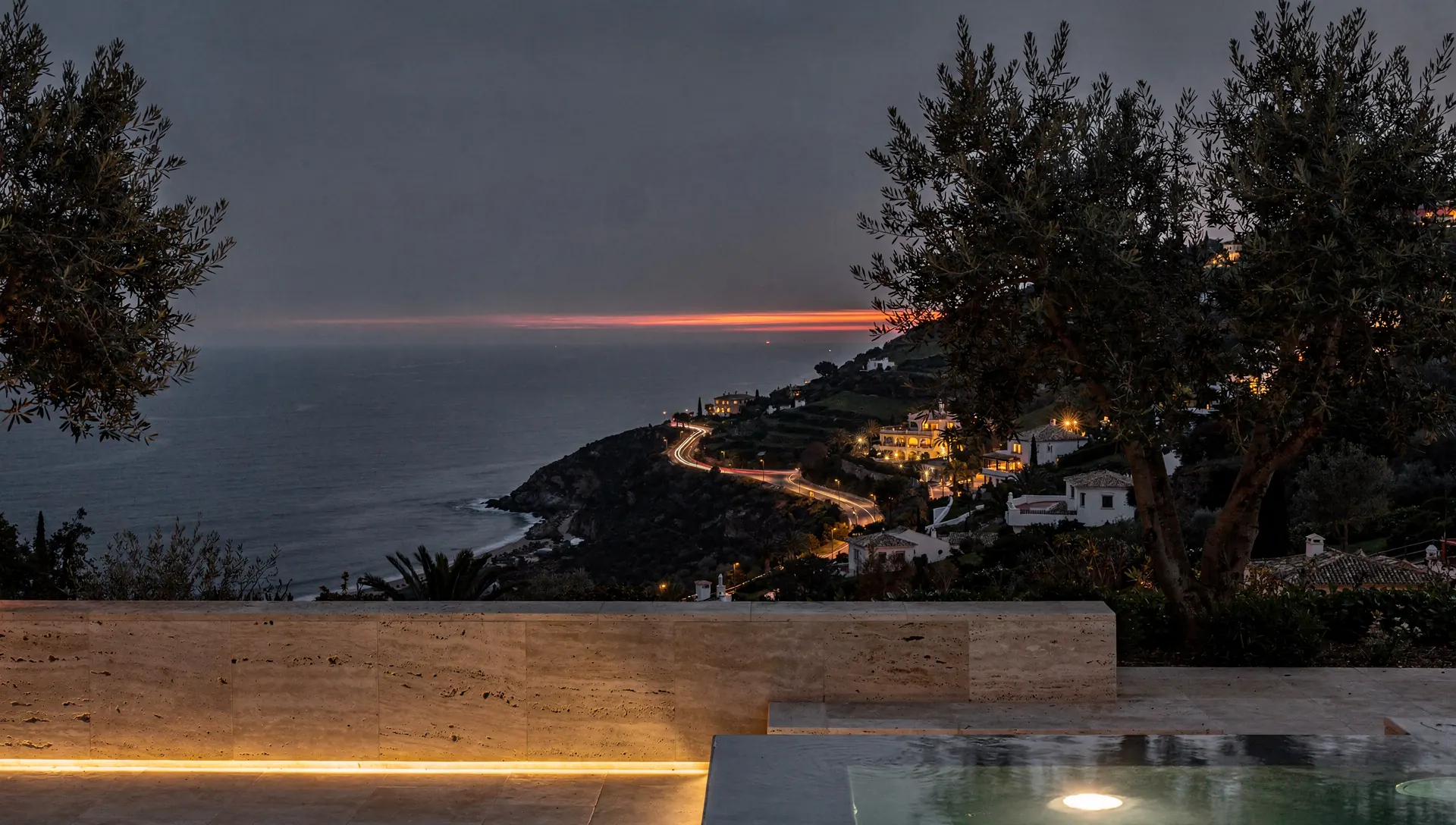 A stone terrace above the coast after sunset, with the lights of the coast road and a thin amber line on the horizon