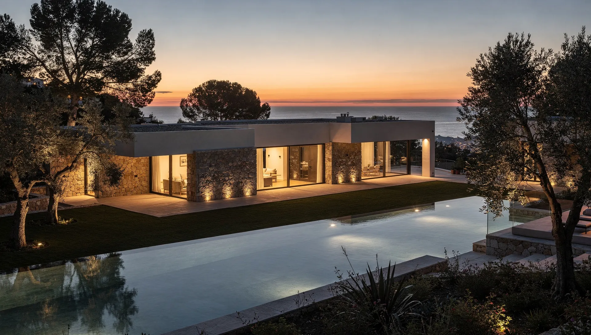 A low contemporary villa with a lit pool above the sea at dusk