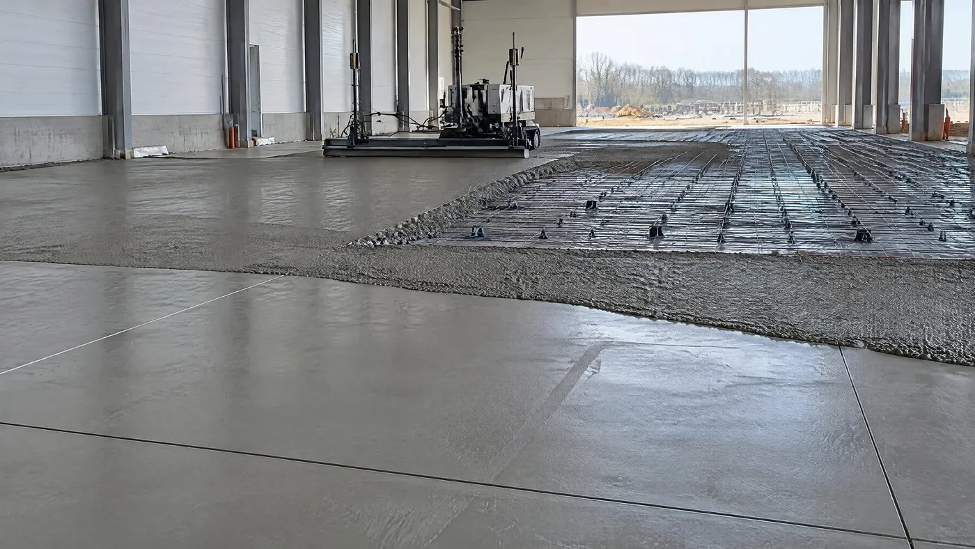 Dalle industrielle en cours de coulage dans un hall&nbsp;: treillis soudé sur cales, béton frais réglé par une règle laser, joints sciés sur la partie durcie