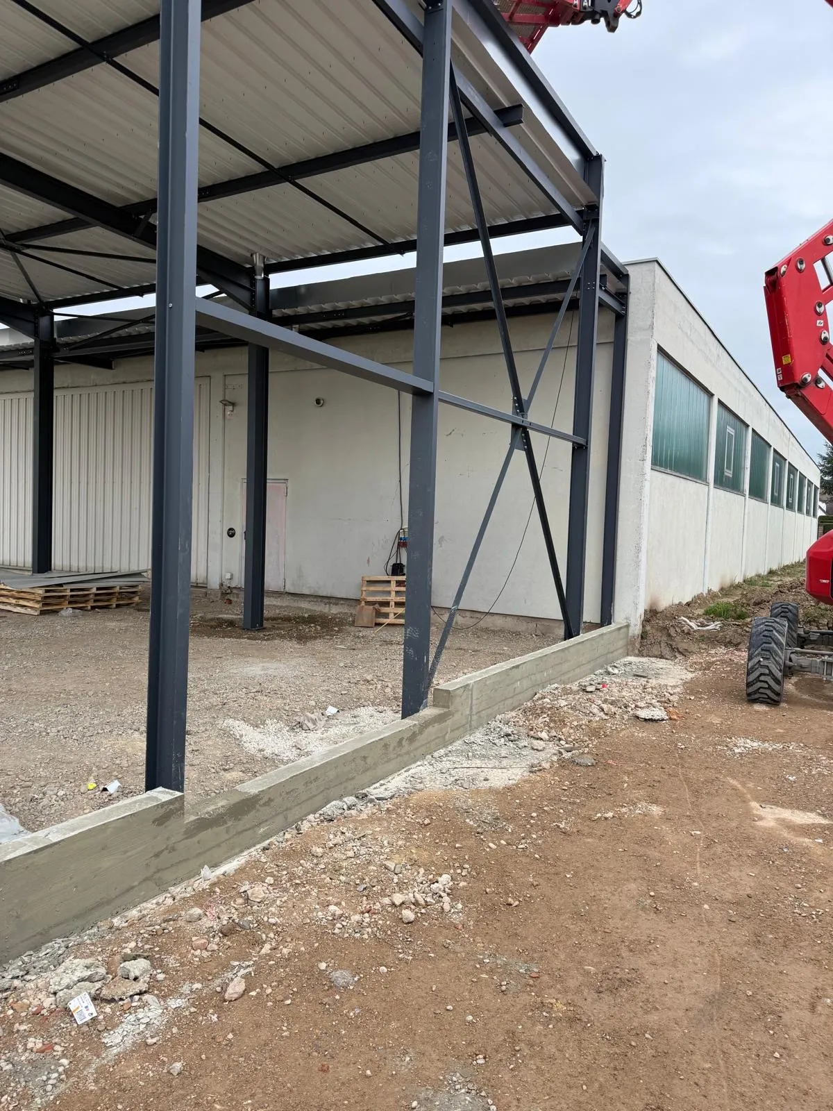 Charpente métallique anthracite d'un hall sur longrines béton fraîches, bâtiment existant en fond