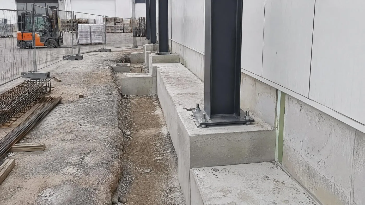 Extension en cours le long d'un hall industriel existant, avec longrines, massifs à platines, joint vertical et premières colonnes métalliques, clôture de chantier devant la zone d'activité