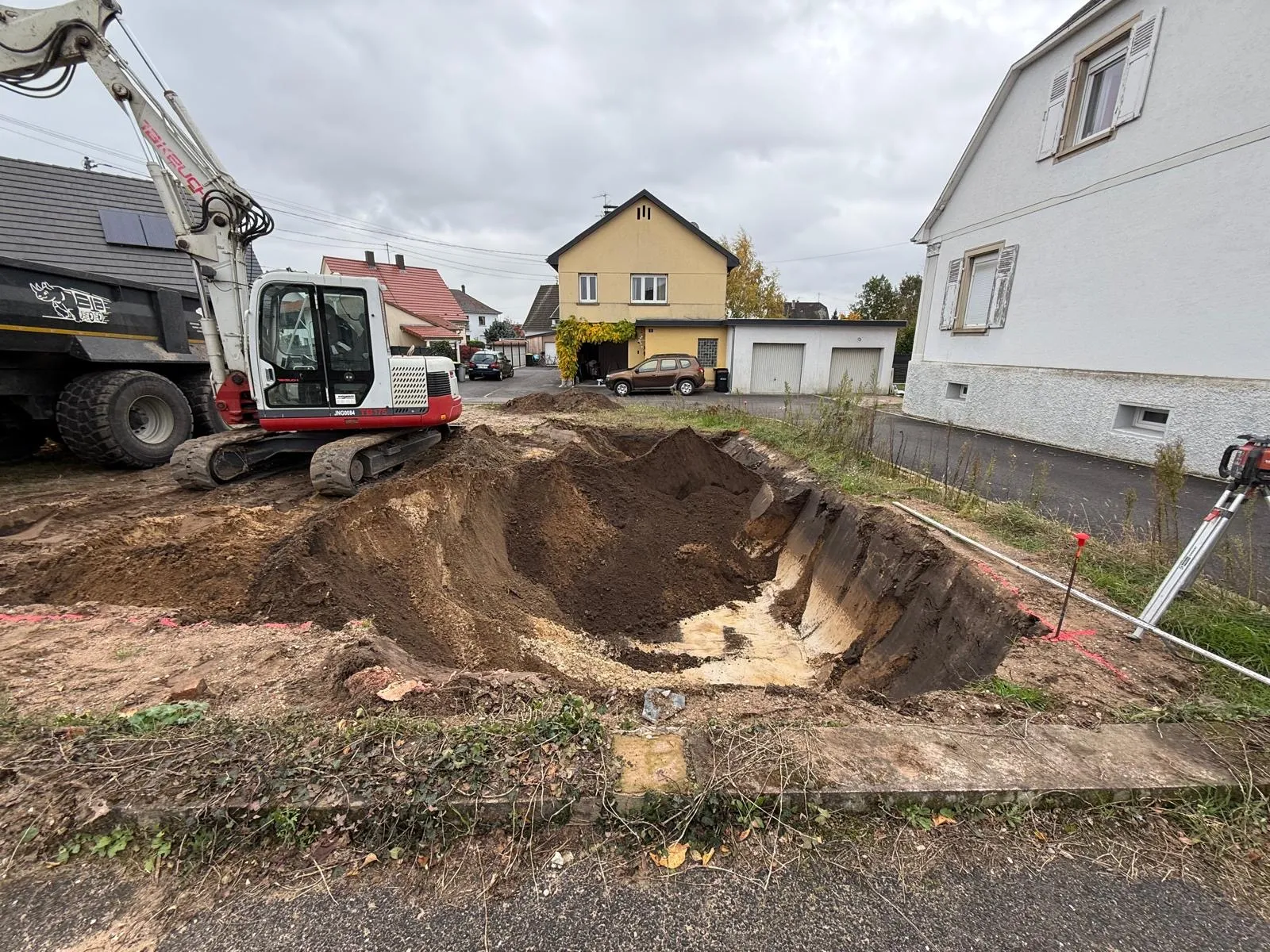 Pelle mécanique ouvrant une fouille sur un terrain à bâtir, maisons voisines en arrière-plan