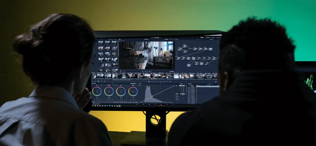 Two creators and contractos sit side by side facing a large monitor, reviewing video editing software with timelines, color grading controls, and node-based adjustments displayed on screen.