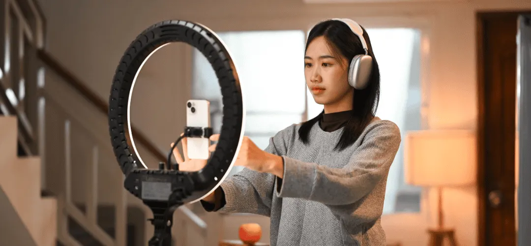 A woman wearing over-ear headphones adjusts a smartphone mounted in front of a lit ring light, preparing to record or stream for her brand deals.