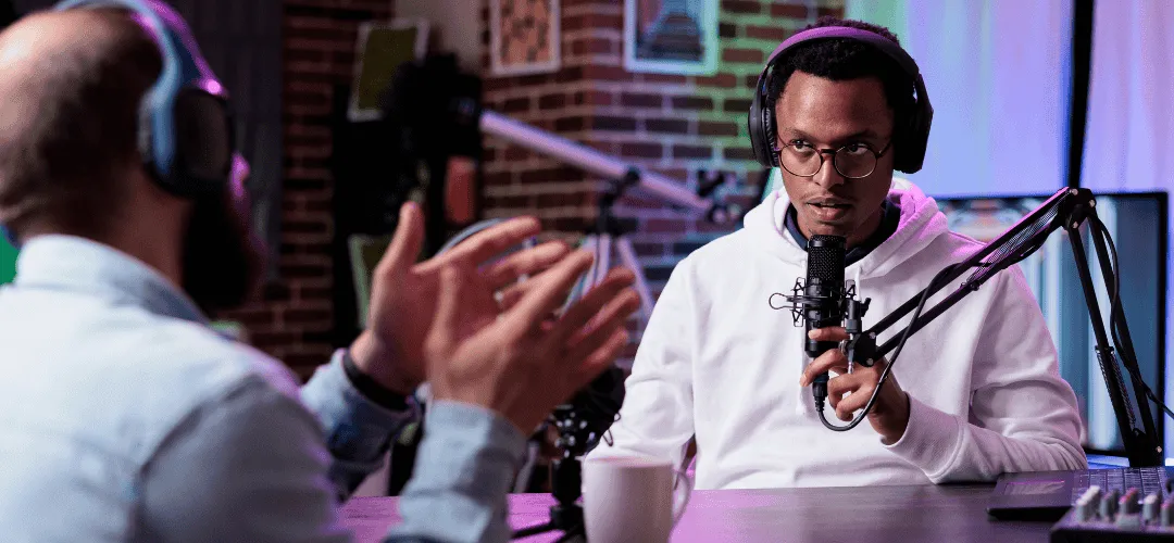 Two men wearing headphones record a podcast in a neon-lit studio, with one speaking into a condenser microphone on a boom arm while the other gestures during conversation.