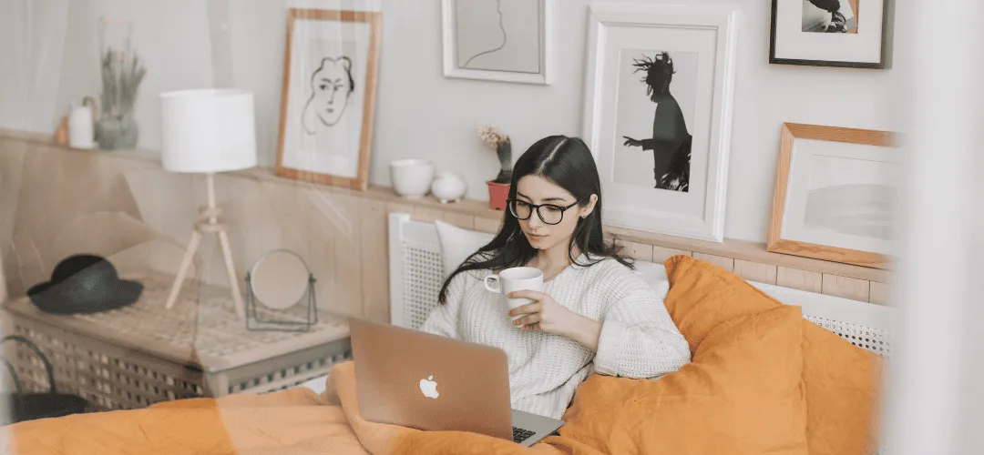 A young woman in glasses and a white sweater works on a MacBook in bed, holding a mug, surrounded by yellow pillows and framed wall art in a cozy bedroom.