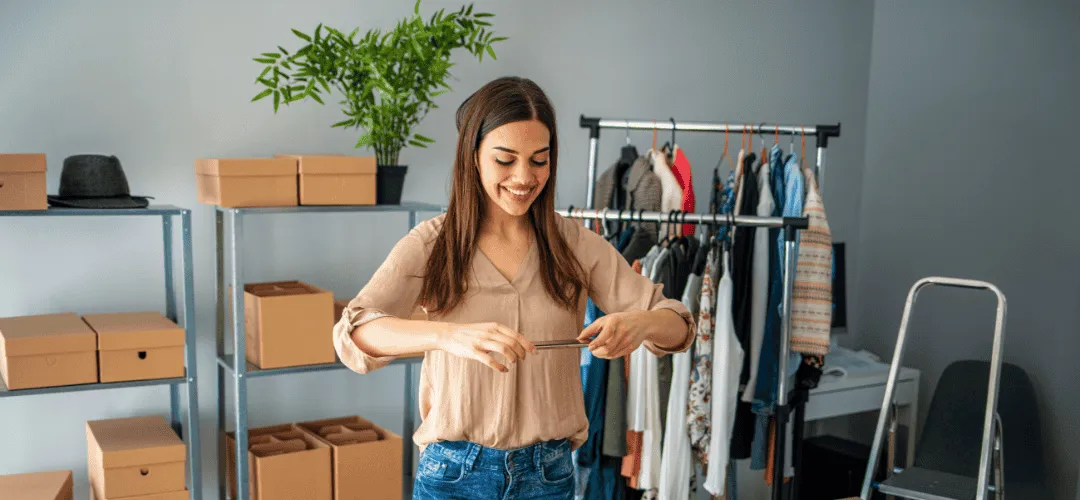 A smiling woman uses her phone to photograph or scan items in a small clothing resale space, surrounded by a garment rack, shelving with cardboard boxes, and a potted plant.