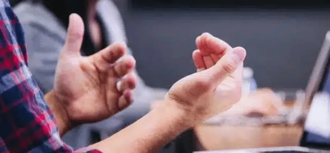 Close-up of two people's hands gesturing during a conversation, with a laptop and blurred figures visible in the background, suggesting a professional consultation or meeting.