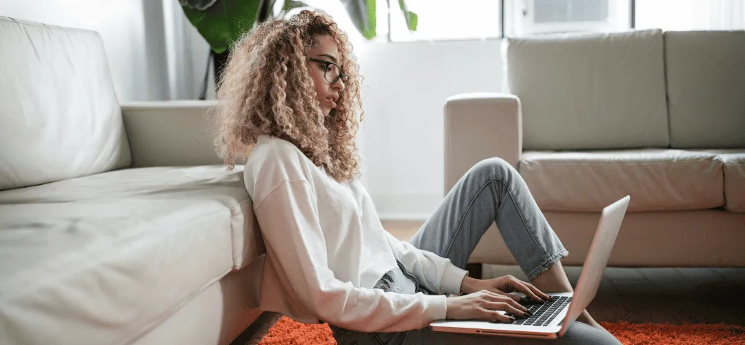 A young woman with curly blonde hair and glasses sits on the floor leaning against a couch, working on a laptop in a bright, casual living room.