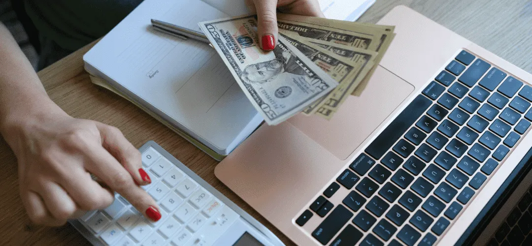 A woman with red nails holds a fan of US dollar bills while using a calculator, with a rose gold laptop and notebook on a wooden desk beside her.