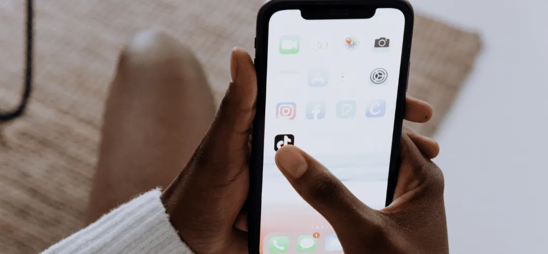 A person taps the TikTok app on an iPhone home screen displaying various social media icons including Instagram and Facebook.