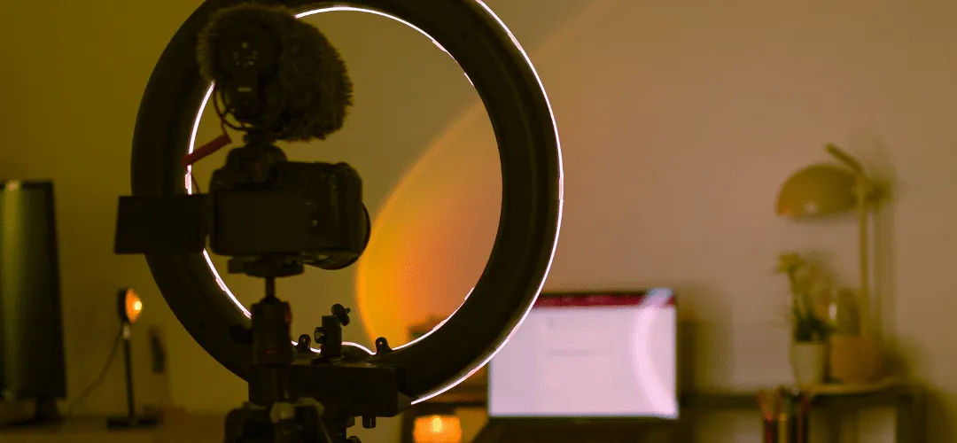 A ring light with a DSLR camera and shotgun microphone mounted on a tripod, glowing with warm amber light against a dimly lit home studio with a monitor and desk lamp in the background.