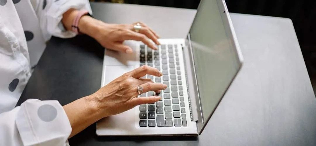 Close-up of a woman's hands typing on a silver laptop on a dark desk, wearing rings and a white polka dot blouse