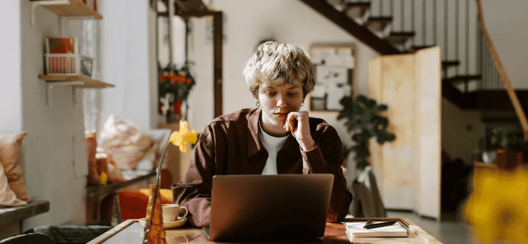 A young person with short blonde hair rests their chin on their hand, focused on a laptop in a warm, eclectic home interior with a staircase and cozy decor in the background.
