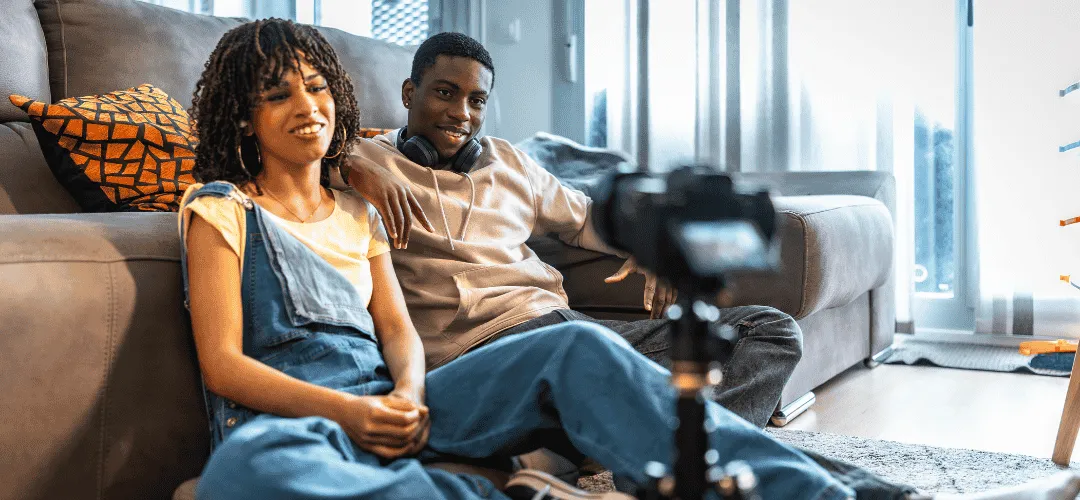 A young man and woman sit casually on the floor against a couch, smiling at a camera on a tripod as they film content together in a bright living room.
