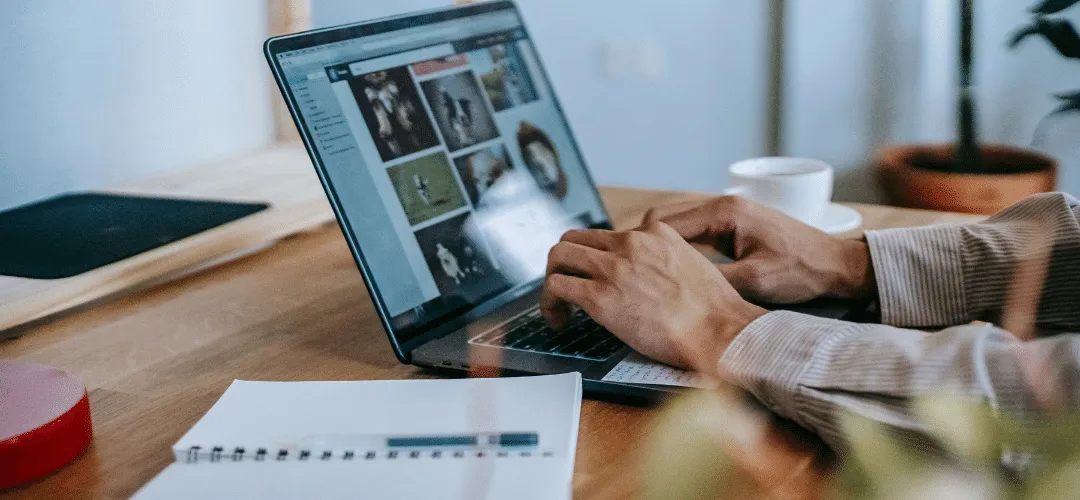 Close-up of hands typing on a laptop displaying a photo editing or media library screen, on a wooden desk with a notebook, coffee cup, and potted plant nearby.