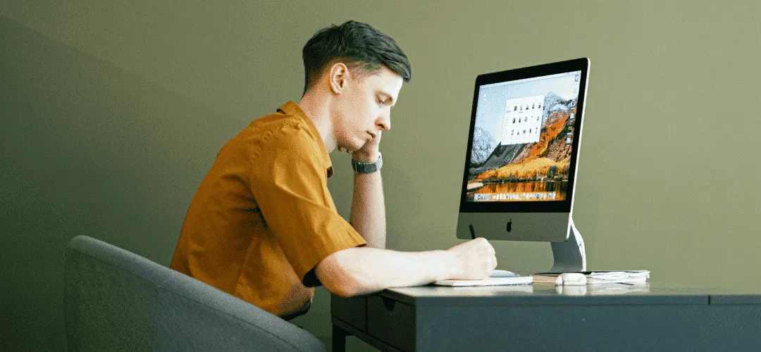 A young man in an orange shirt rests his chin on his hand, staring pensively at an iMac on a minimal desk against an olive green wall.