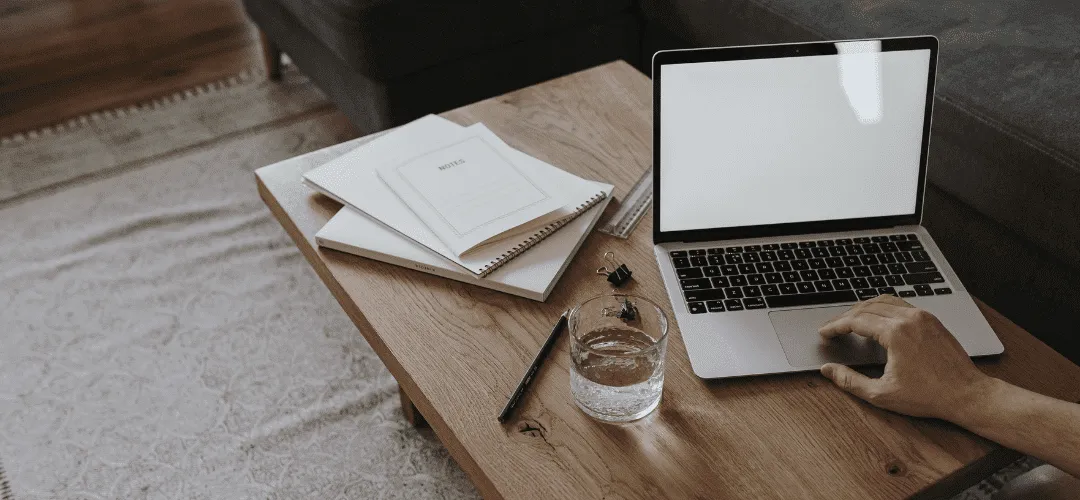 A hand uses a laptop on a rustic wooden coffee table alongside stacked notebooks, a pen, binder clips, and a glass of water, with a couch and rug in the background.