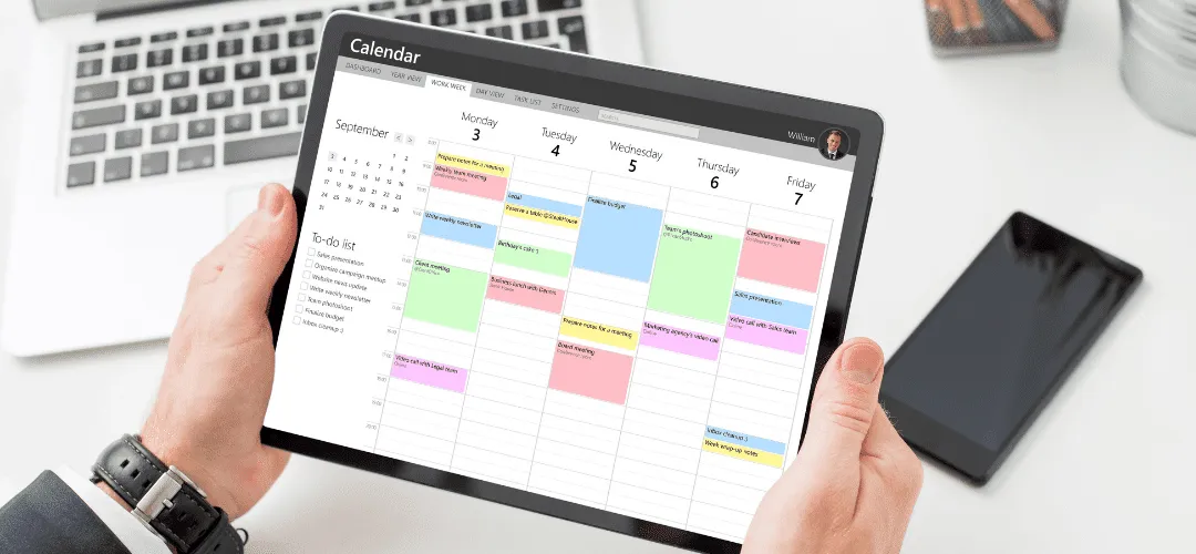 A man holds a tablet displaying a colorful weekly calendar app for September, with a laptop, smartphone, and business card holder visible on the desk behind it.