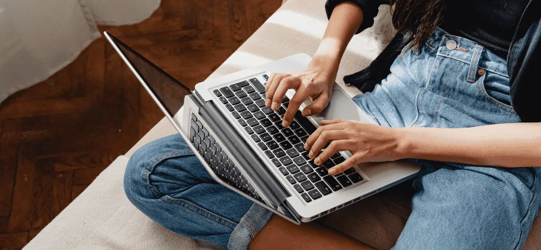 Overhead view of a woman in jeans sitting cross-legged on the floor, typing on a laptop balanced on her knees over a white rug and hardwood floor.
