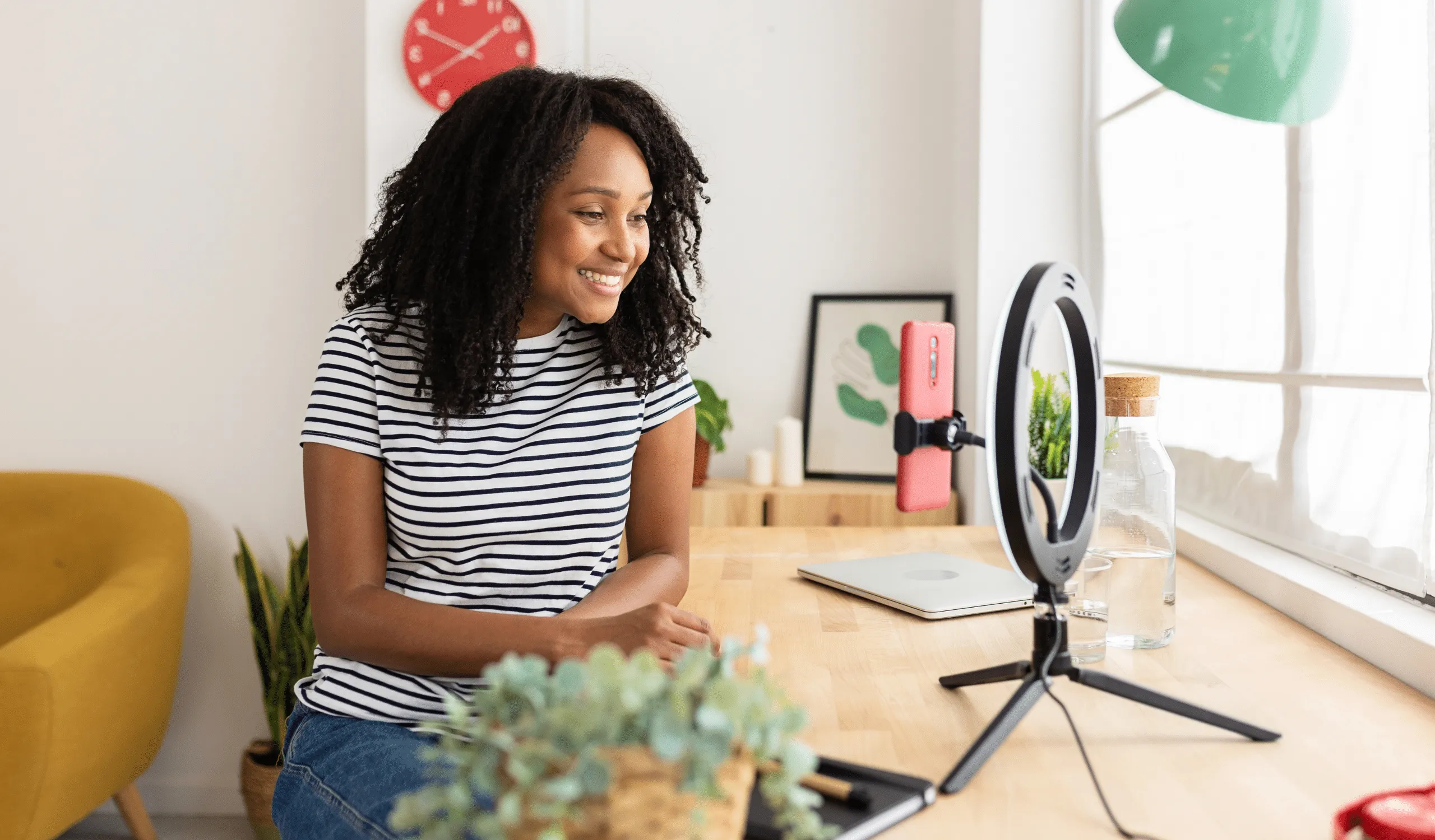 A smiling woman in a striped t-shirt records content at a bright home desk, with a phone mounted on a ring light, a laptop, and a red clock on the wall behind her.
