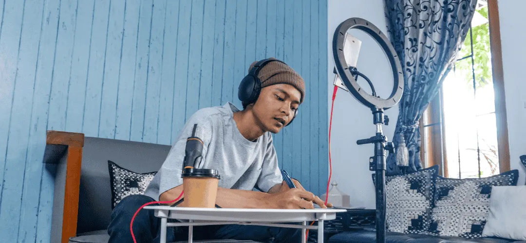 A young man in a beanie and headphones writes notes at a small table beside a ring light and microphone, set up for content creation in a blue walled room.