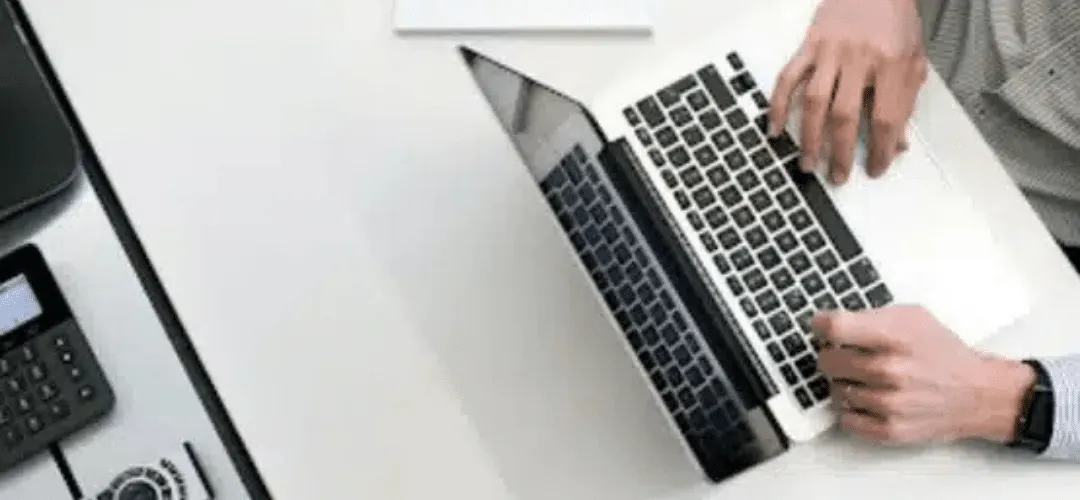 Overhead view of a man's hands typing on a laptop on a white desk, with a desk phone visible to the left.