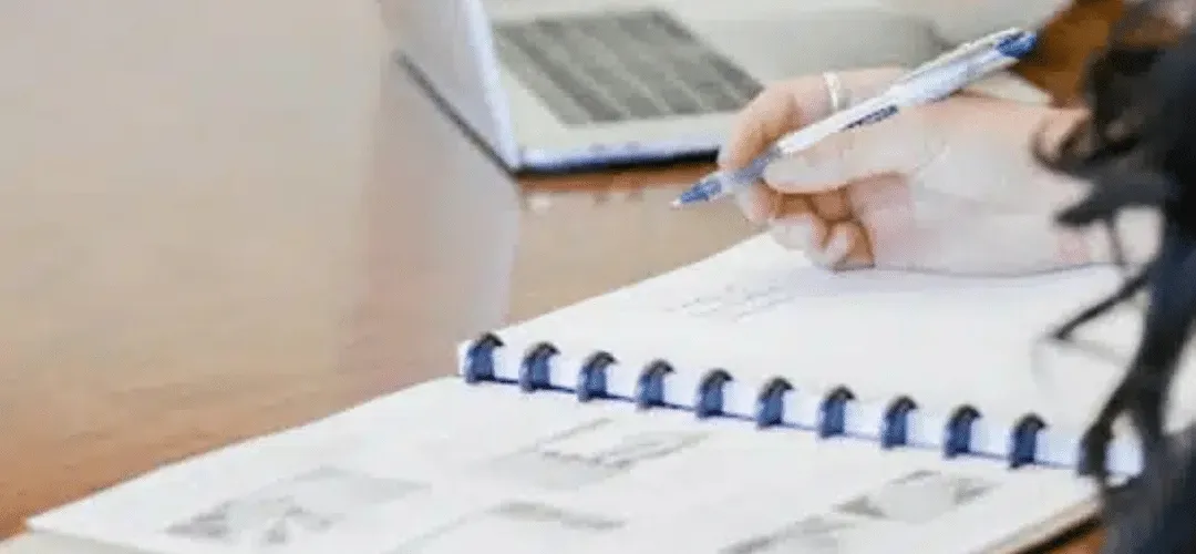 Close-up of a woman's hand writing in a spiral notebook with a pen, with a laptop blurred in the background on a wooden desk.